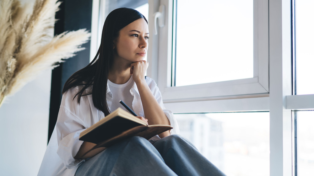 Thoughtful female sitting on windowsill with notebook